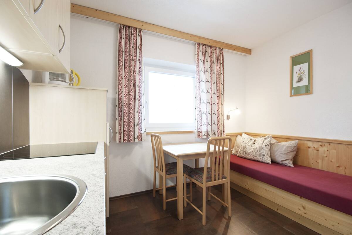 A compact kitchen with a dining area, a wooden bench with cushions, and a window with patterned curtains.