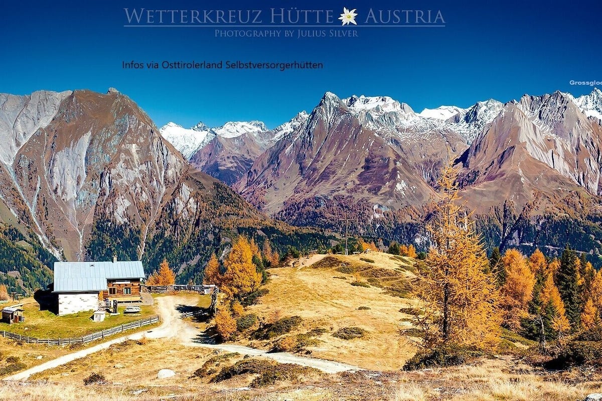 A mountain lodge with a wooden cabin, surrounded by autumn-colored trees and snow-capped mountains.