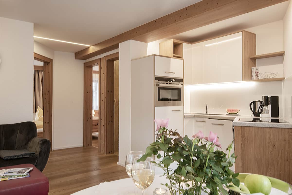 A modern kitchen with white cabinets, a built-in oven, and a coffee maker. There is a dining area with a red sofa and a table with a vase of flowers.