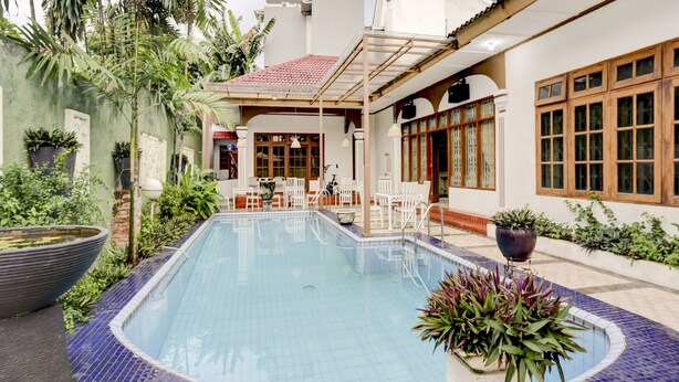 A swimming pool with a tiled edge, surrounded by potted plants and a seating area with white chairs and tables. - Capital O 90066 Tobana Pejaten House (Jakarta)