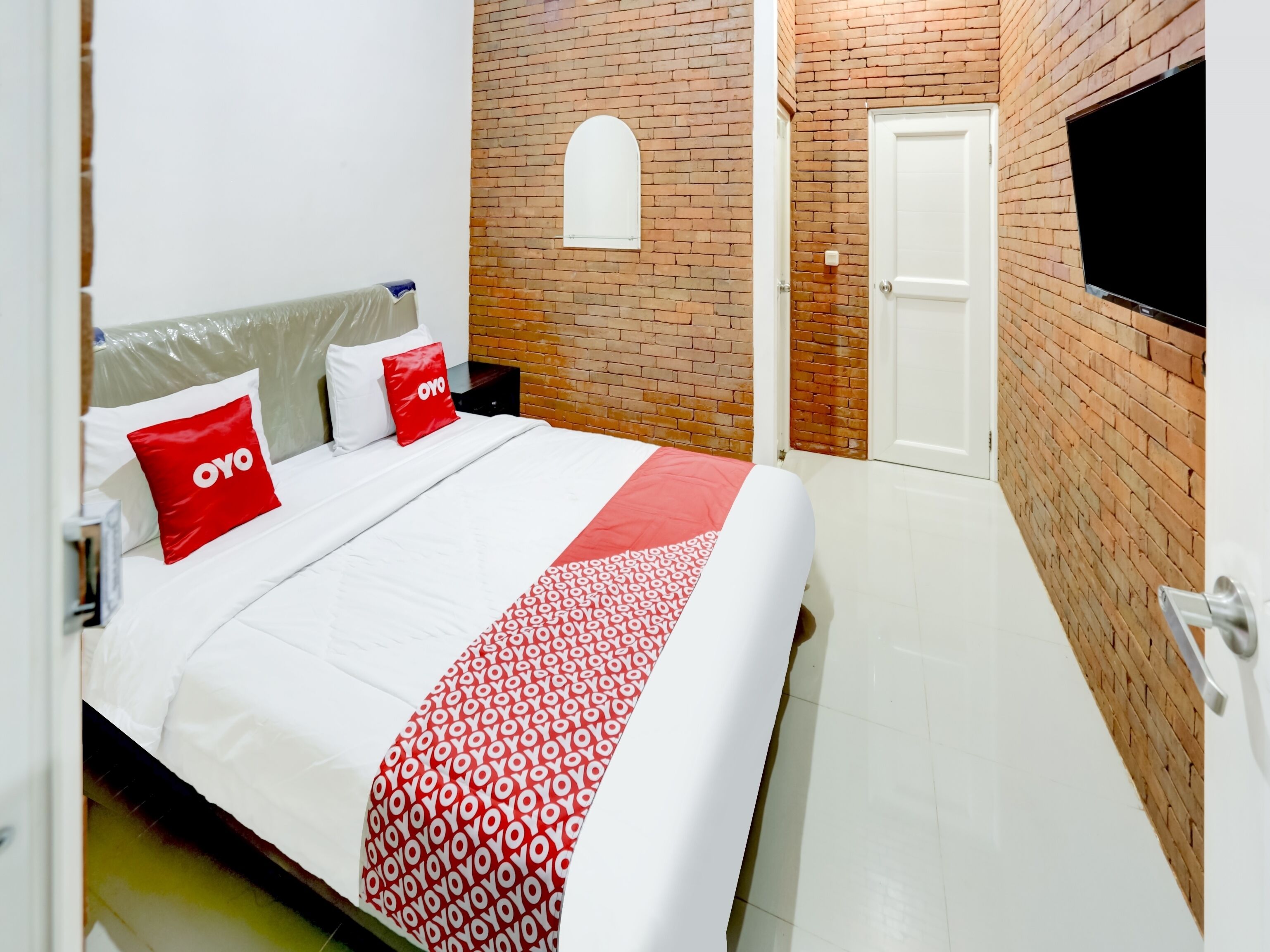 Une chambre d’hôtel avec un mur en briques, un téléviseur fixé au mur, un lit avec une literie blanche et des oreillers rouges, et un plaid à motifs.