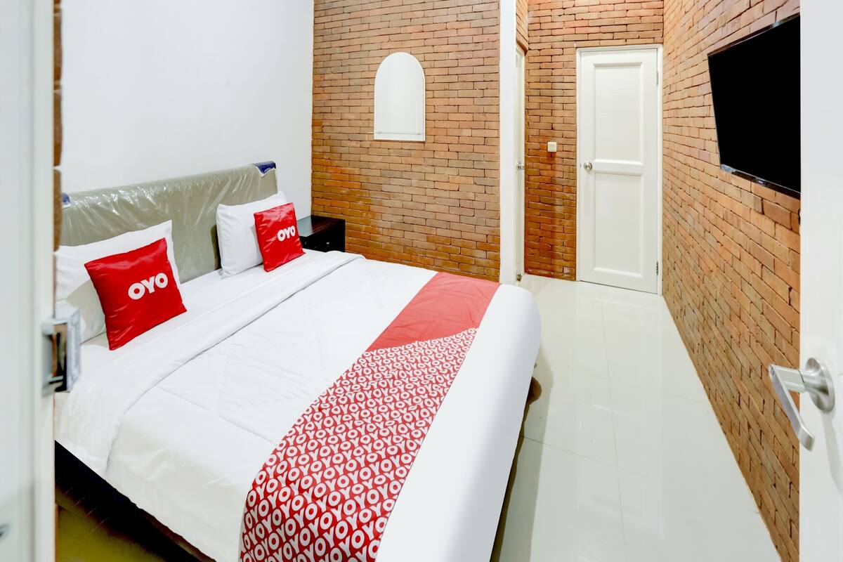 Ein Hotelzimmer mit Backsteinwand, einem an der Wand montierten Fernseher, einem Bett mit weißer Bettwäsche und roten Kissen sowie einer weißen Tür.