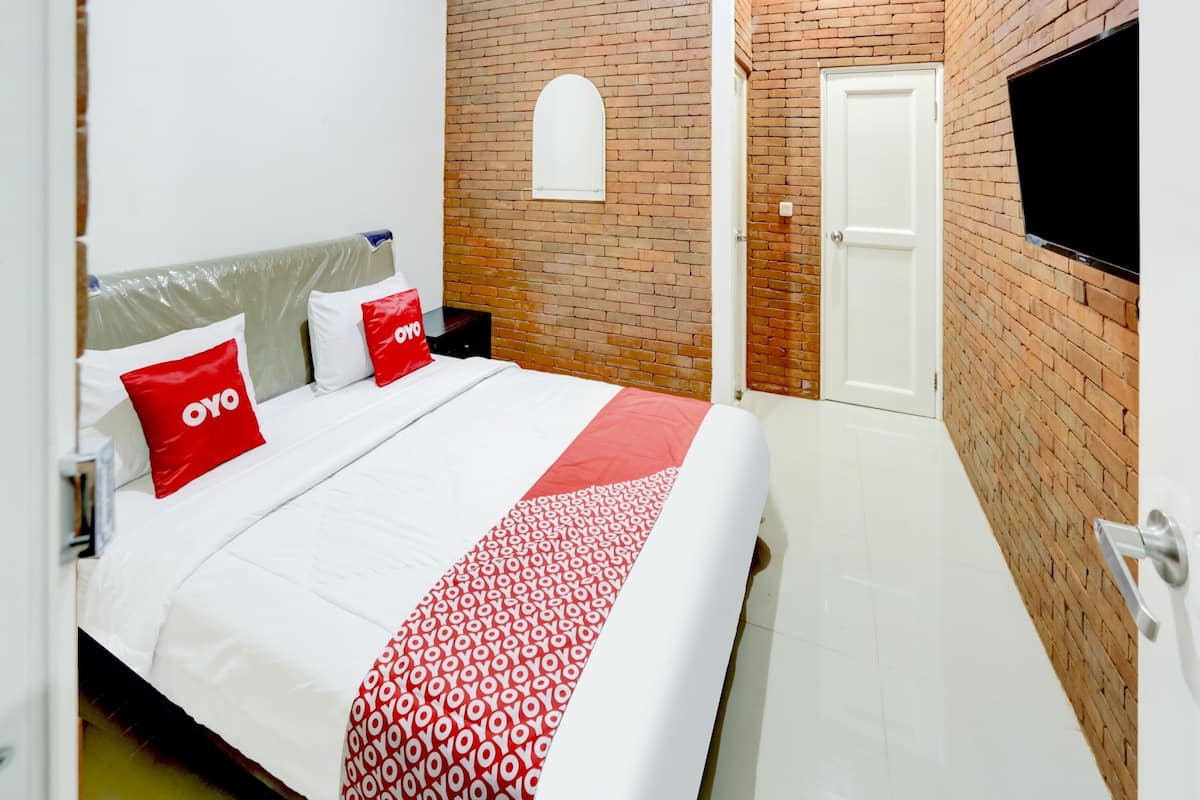 Une chambre d’hôtel avec un mur en briques, un téléviseur fixé au mur, un lit avec une literie blanche et des oreillers rouges, et un plaid à motifs.