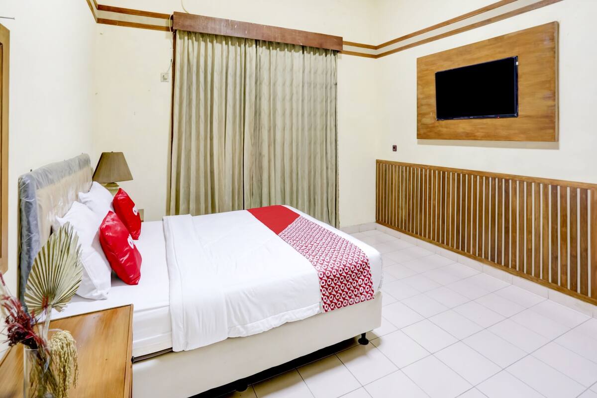 Ein ordentlich eingerichtetes Hotelzimmer mit Bett, Nachttischlampe, einem an der Wand befestigten Fernseher und einem großen Fenster mit Vorhängen.