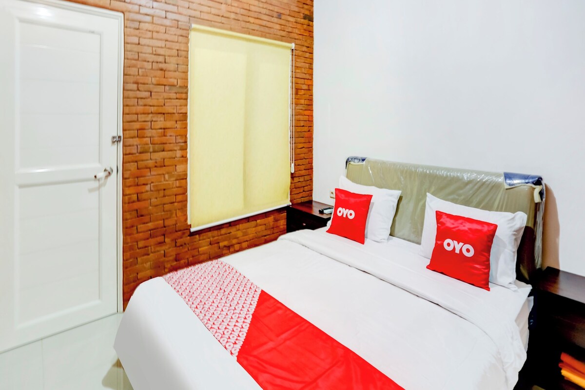 A hotel room with a brick wall, a white door, a bed with red and white bedding, and a window with a yellow blind.