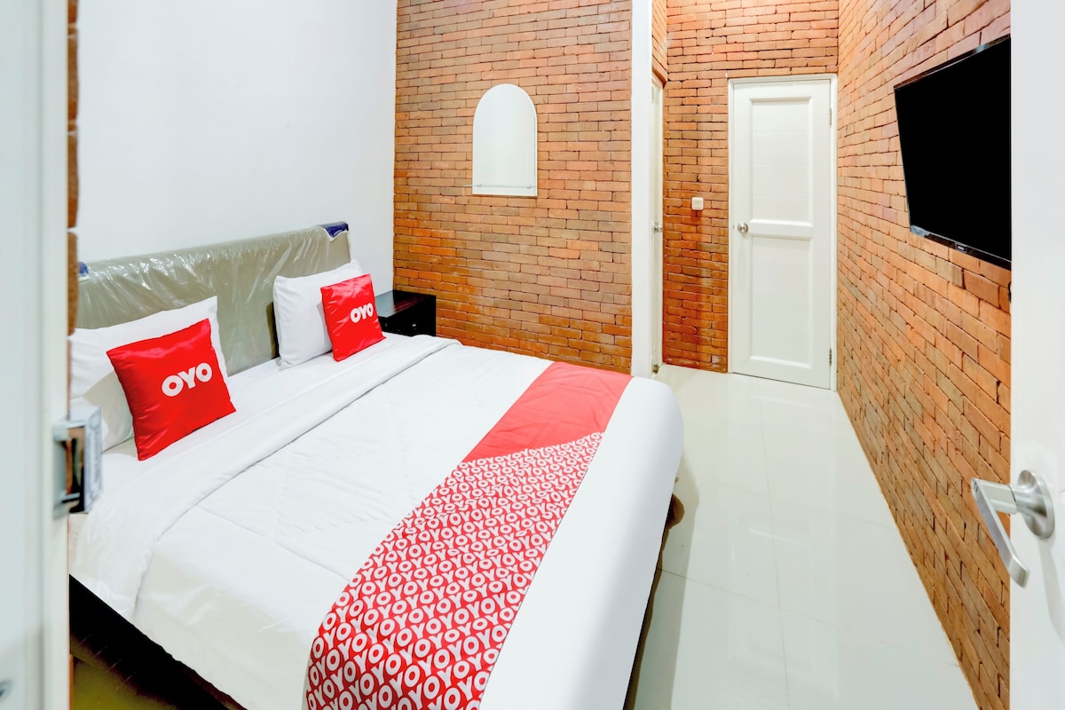 A hotel room with a brick wall, a TV mounted on the wall, a bed with white bedding and red pillows, and a white door.
