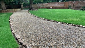 A gravel pathway leading to a grassy area with a brick wall and trees in the background. - Piper Cottage, Bilbrough. (York)