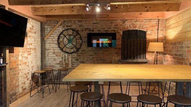 A rustic interior with a wooden bar table, stools, a large clock, a fireplace, and a television. - Piper Cottage, Bilbrough. (York)