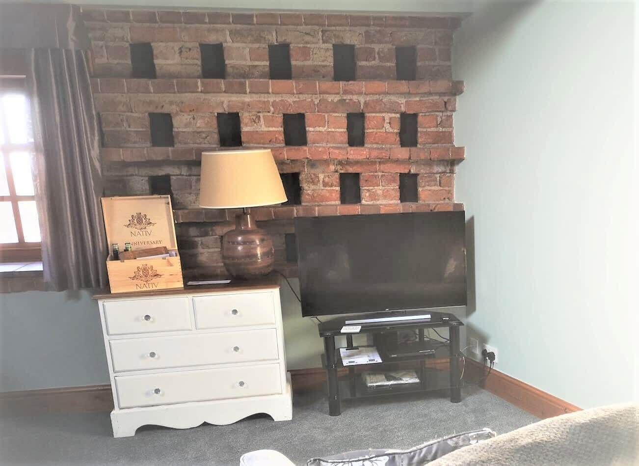 A room with a brick fireplace, a television on a stand, a white dresser with drawers, and a lamp.