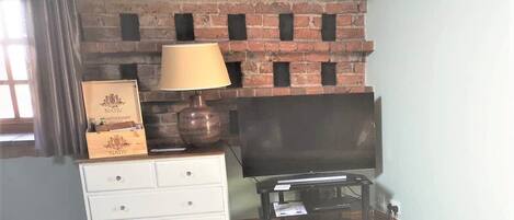 A room with a brick fireplace, a television on a stand, a white dresser with drawers, and a lamp.