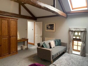 A cozy room with a wooden wardrobe, a sofa with patterned cushions, a small table, and a window with curtains. - The Carriage House, Bilbrough, York. (York)
