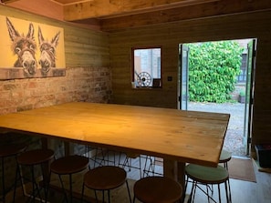 Interior de fusta amb un gran taulell i quatre tamborets. Hi ha una paret de maó amb un mural d'asees, una finestra amb un rellotge i vista a un jardí verd.