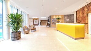 Un hall d’hôtel moderne avec un comptoir d’accueil jaune vif, une grande plante en pot et un espace salon spacieux offrant une vue sur la ville.