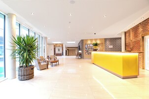 Um saguão de hotel moderno com uma mesa de recepção amarelo-vivo, uma grande planta em vaso e uma área de estar espaçosa com vista para a cidade.