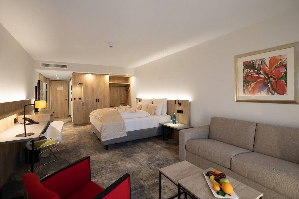 Une chambre d’hôtel avec un lit, un canapé, un bureau avec une chaise, une petite table avec des fruits et un tableau au mur.