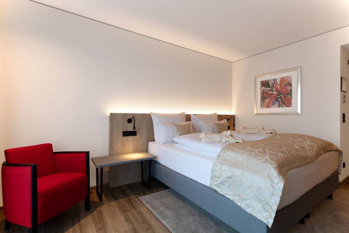 Une chambre d’hôtel moderne avec un lit, une chaise rouge, une table de chevet et une lampe fixée au mur.