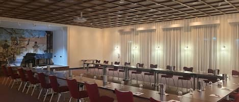 Un salón de conferencias con una larga mesa, sillas rojas y un mural en la pared de la izquierda.