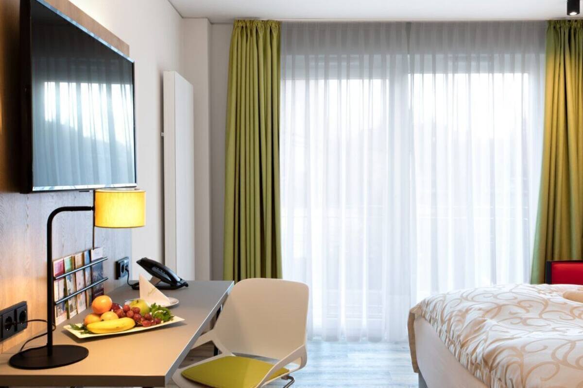 Une chambre d’hôtel avec un lit, un bureau avec une lampe, une chaise, une télévision et une fenêtre avec des rideaux.