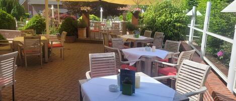 Een terras buiten met tafels en stoelen, overdekt met grote parasols, omringd door groen en bakstenen muren.