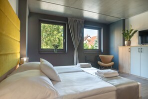 A modern bedroom with a bed, a chair, a TV, and a window with a view of trees and buildings.