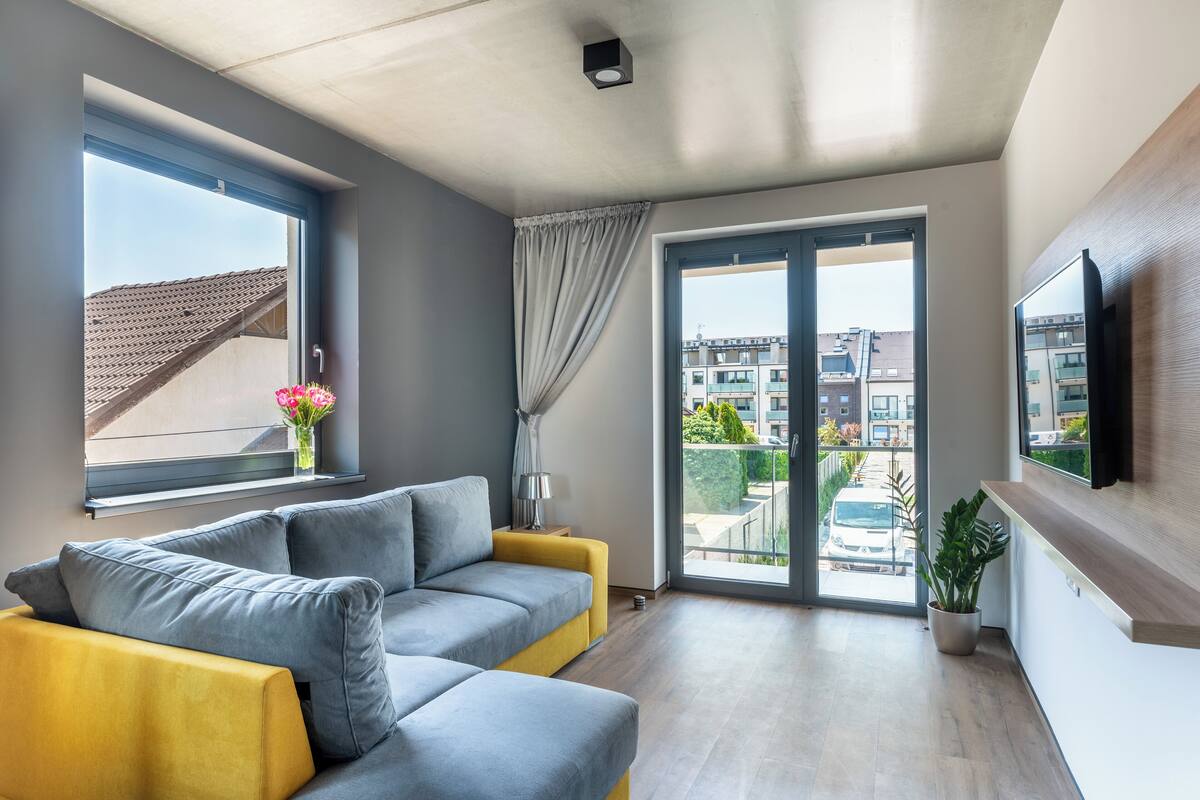 Una sala de estar moderna con un sofá gris y amarillo, un televisor montado en la pared y un amplio ventanal corredero que da a un balcón exterior.