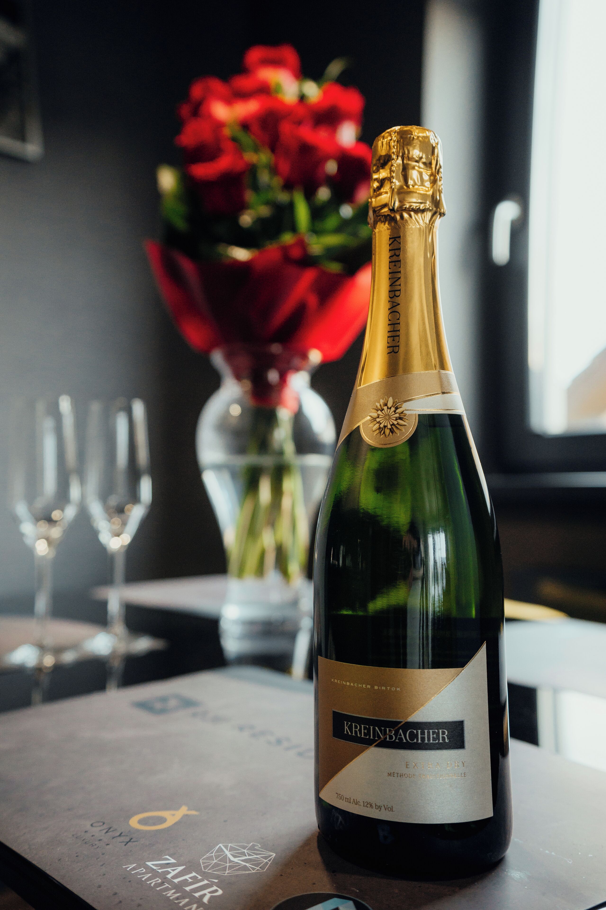 Eine Flasche Kreinbacher Champagner auf einem Tisch, im Hintergrund eine Vase mit roten Rosen und zwei leere Gläser.