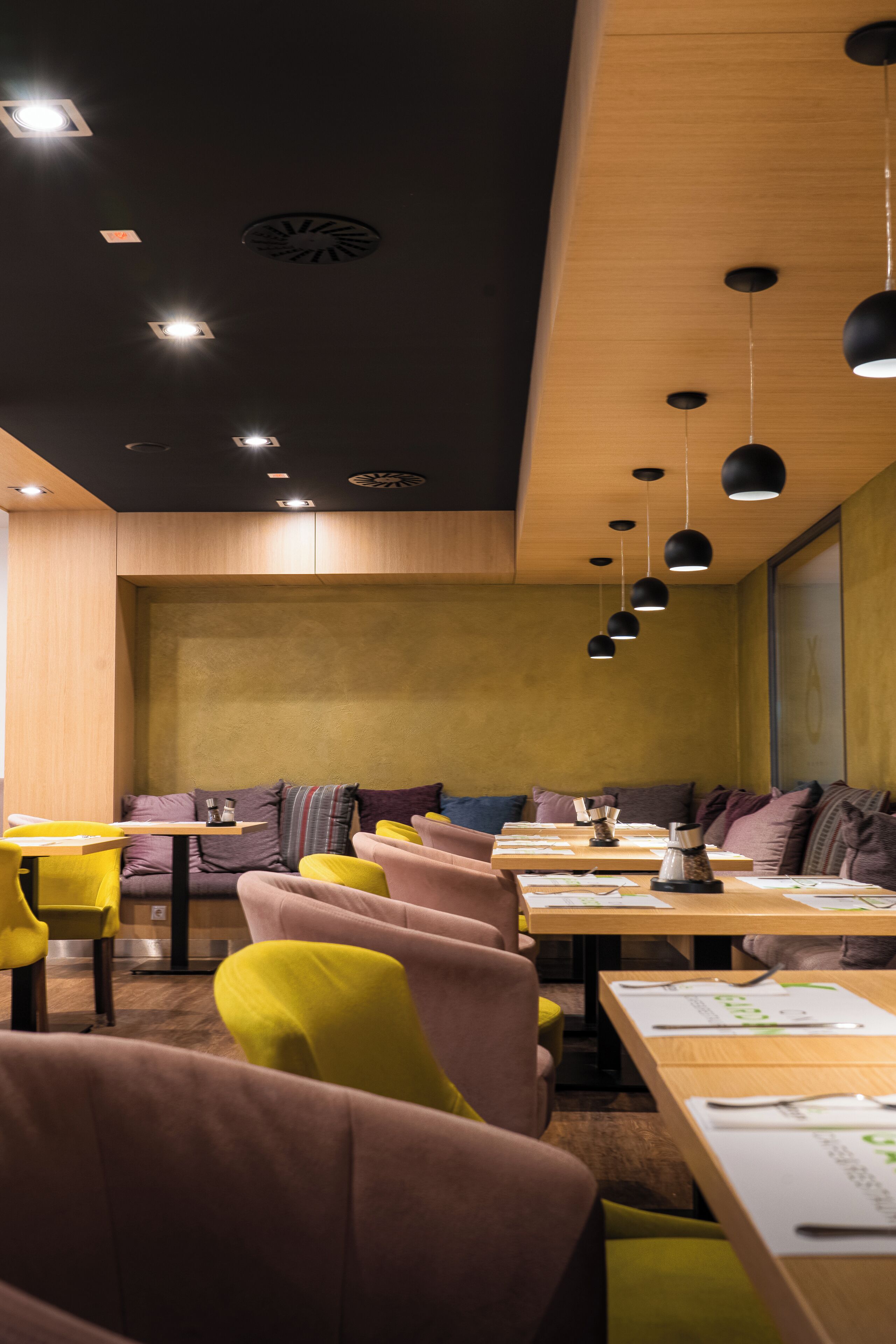 L’intérieur d’un restaurant moderne, avec des chaises jaunes et roses, des tables en bois et des suspensions noires.