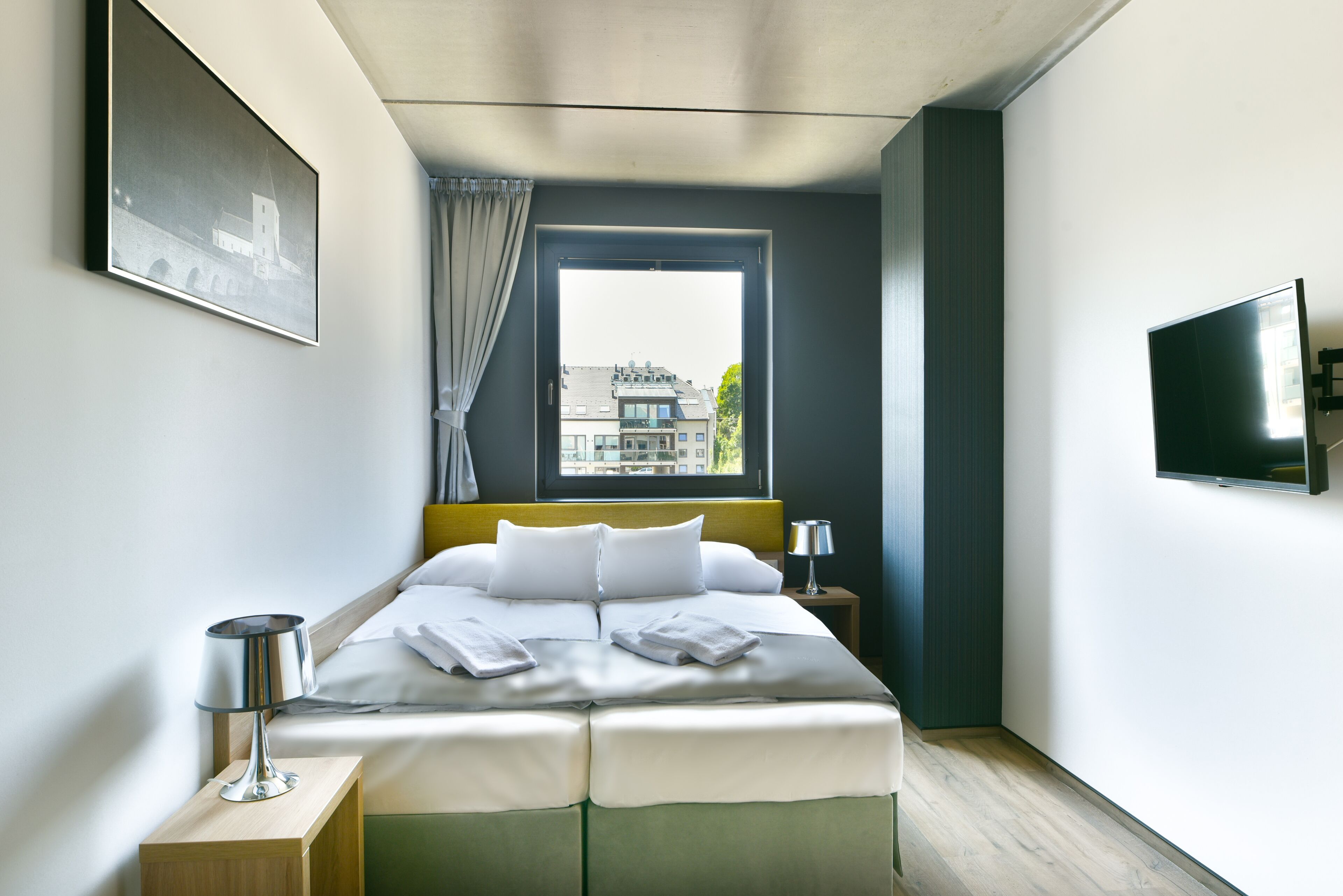 Een moderne hotelkamer met een net opgemaakt bed, een aan de muur bevestigde televisie en een raam met uitzicht op gebouwen.