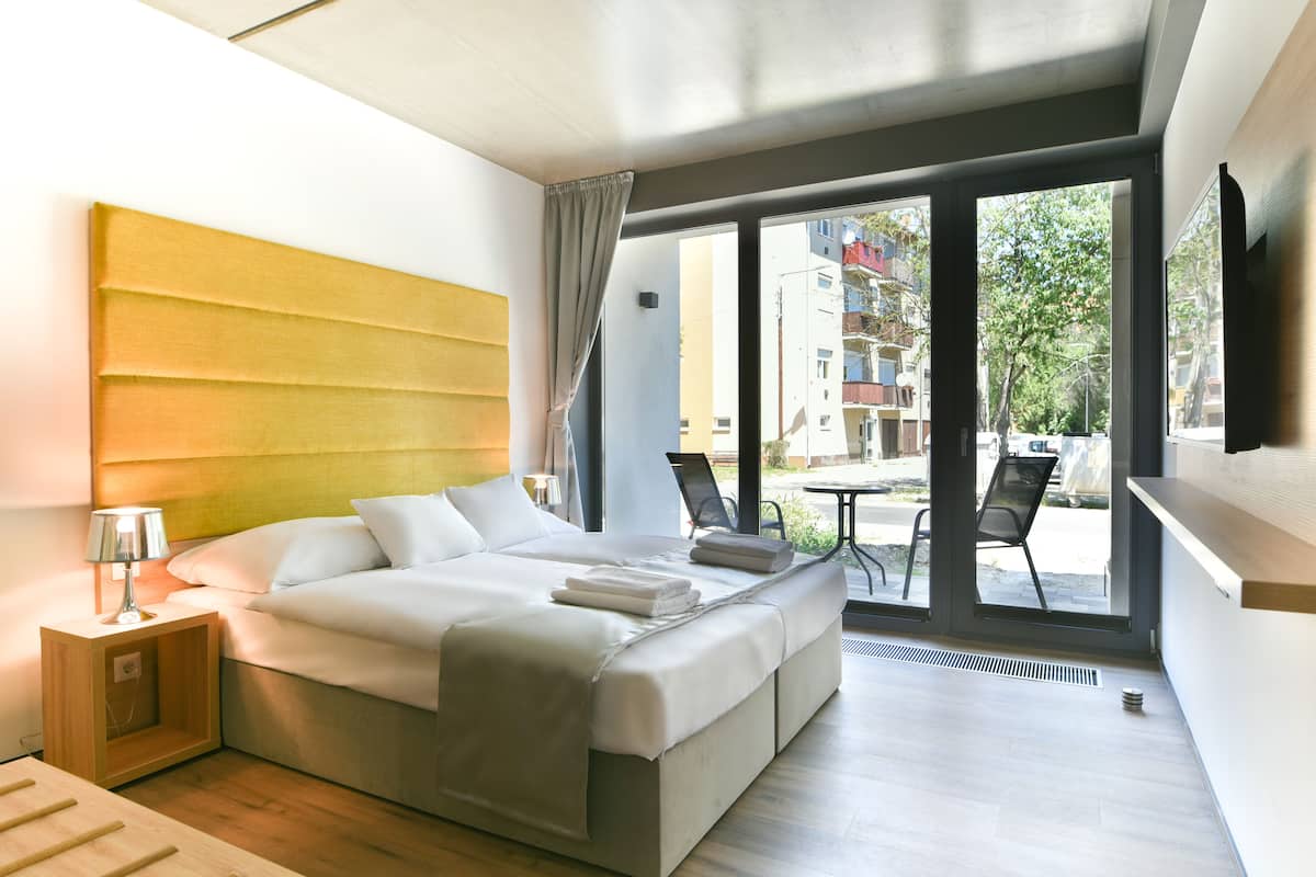 Habitación de hotel moderna con una cama grande, una mesita de noche con lámpara, vistas al exterior a través de amplios ventanales y una pequeña zona de balcón.