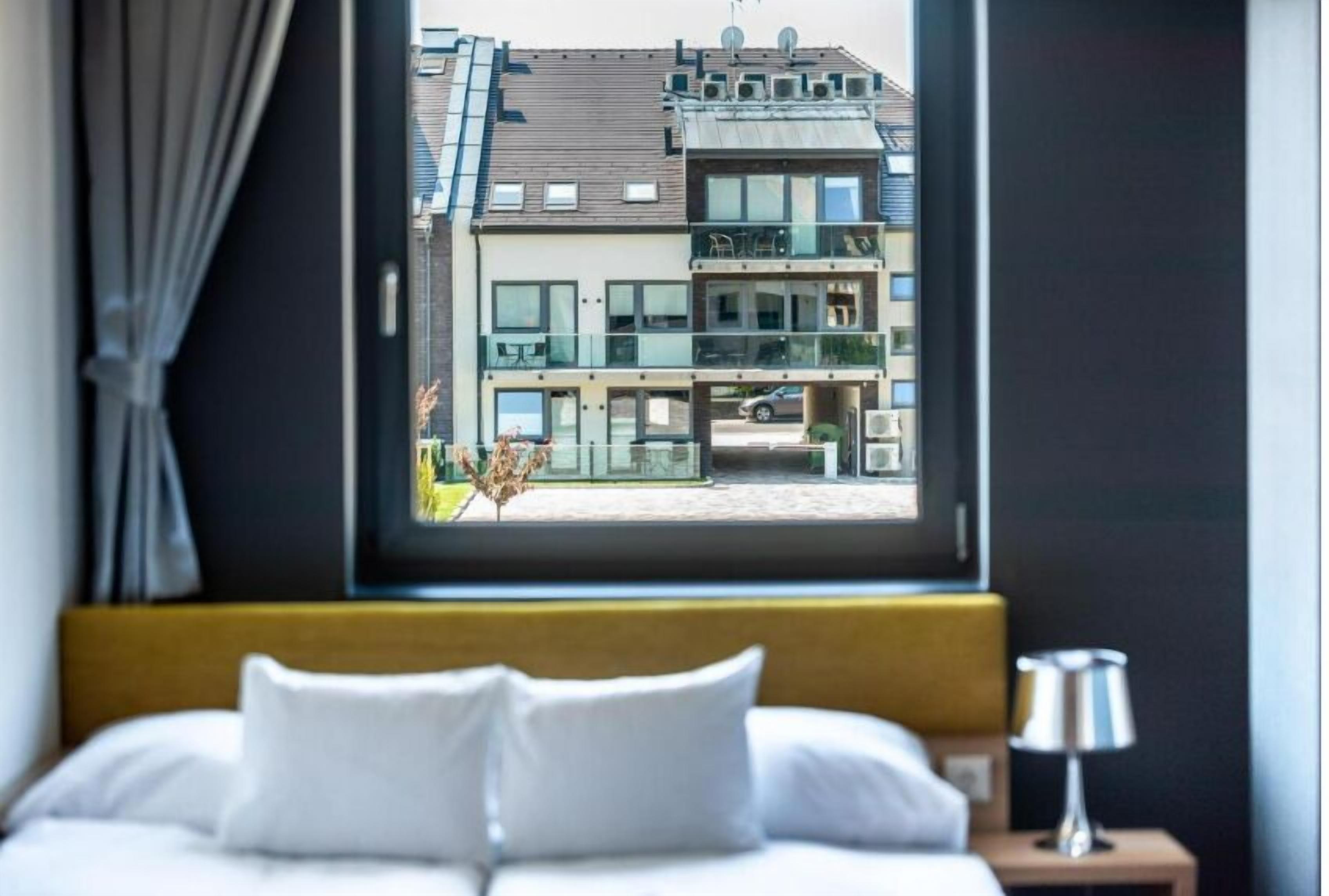 A hotel room with a bed, a window with a view of a modern building, and a bedside table with a lamp.