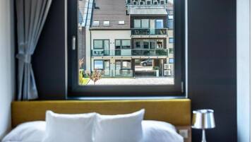 A hotel room with a bed, a window with a view of a modern building, and a bedside table with a lamp.