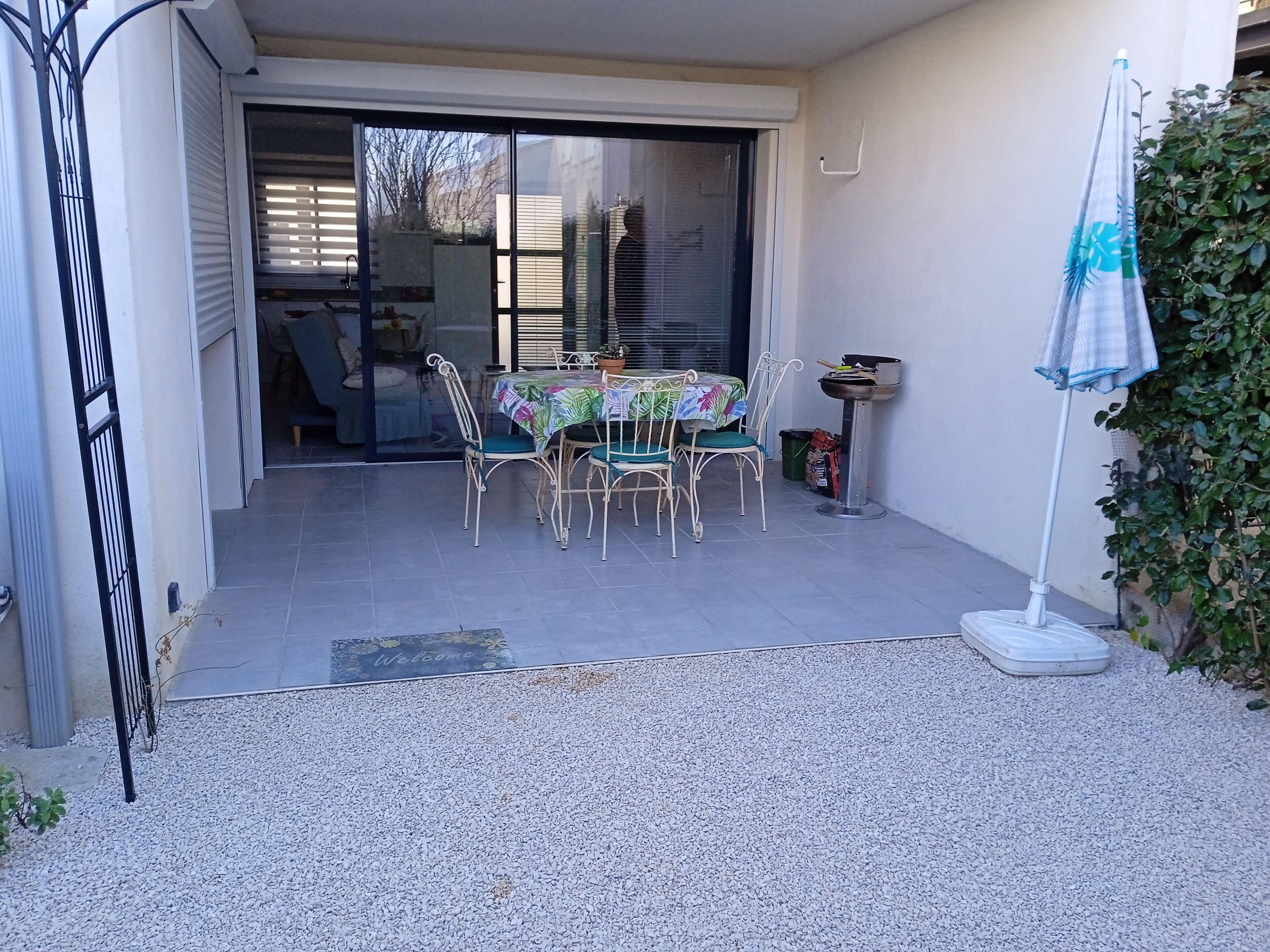 Un patio con mesa y sillas, un pequeño jardín y un ventanal corredera que da a un espacio interior.
