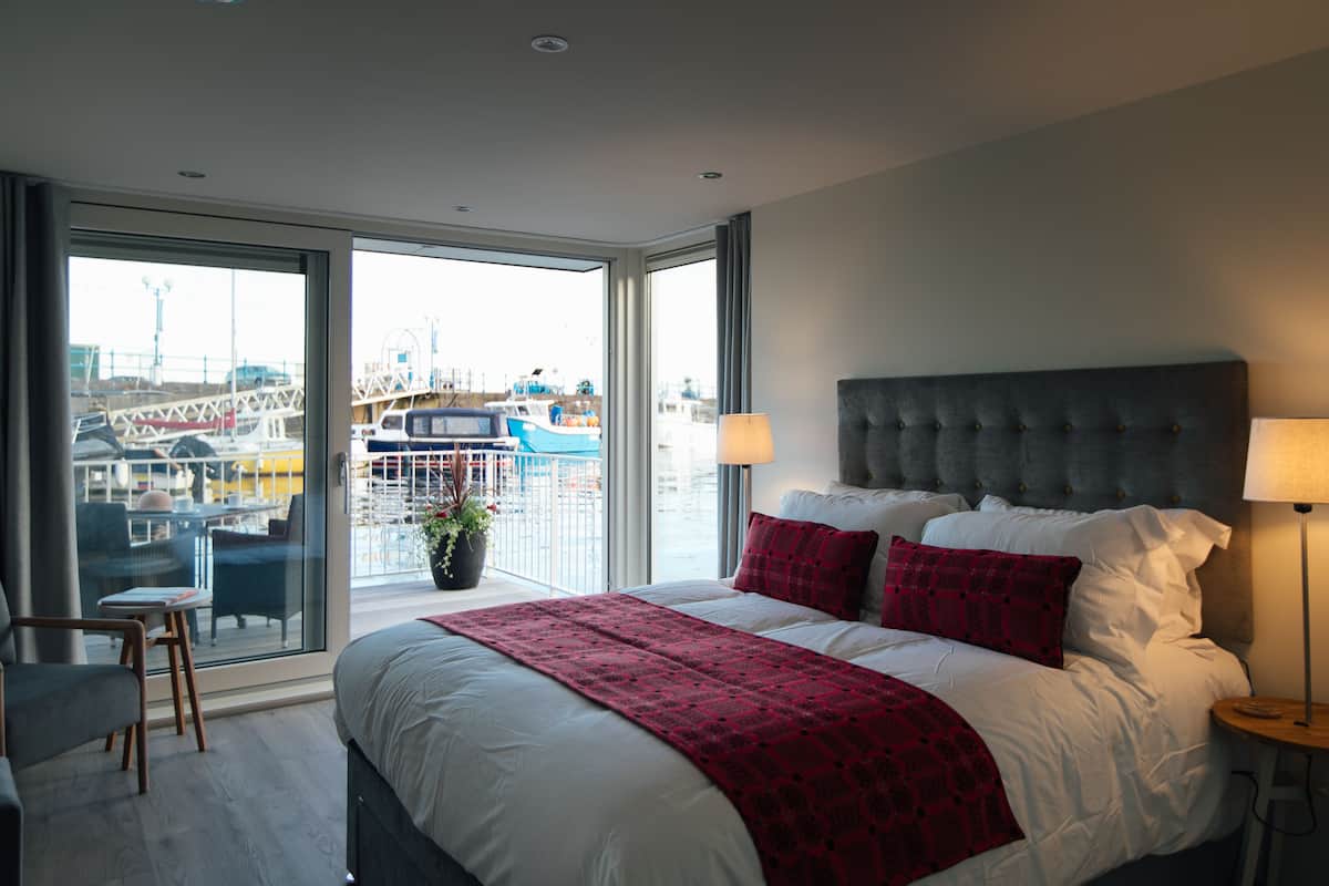 Ein modernes Hotelzimmer mit einem großen Bett, zwei Nachttischlampen und Blick auf eine Marina durch raumhohe Fenster.