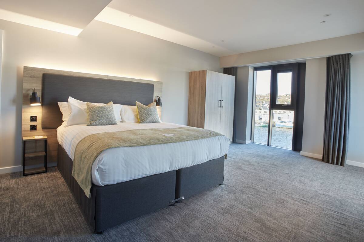 Ein modernes Hotelzimmer mit einem großen Bett, einem Nachttisch, einem Kleiderschrank und Blick ins Freie durch das Fenster.