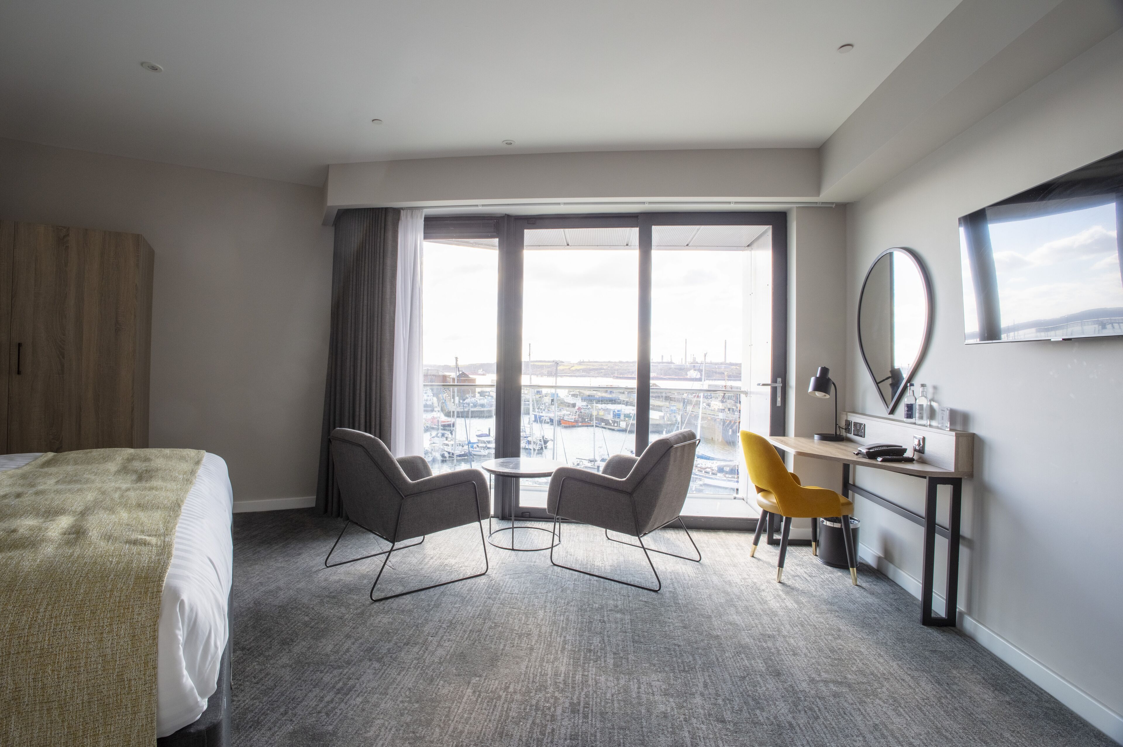 A hotel room with a bed, two chairs, a desk, a large window with a view of the city, and a mirror on the wall.