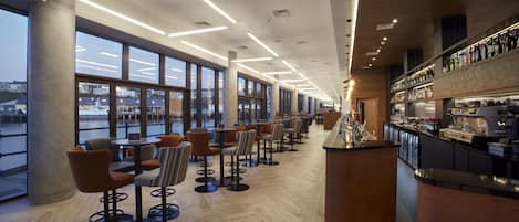 A modern bar area with seating and a bar counter, large windows, and a view of the waterfront.