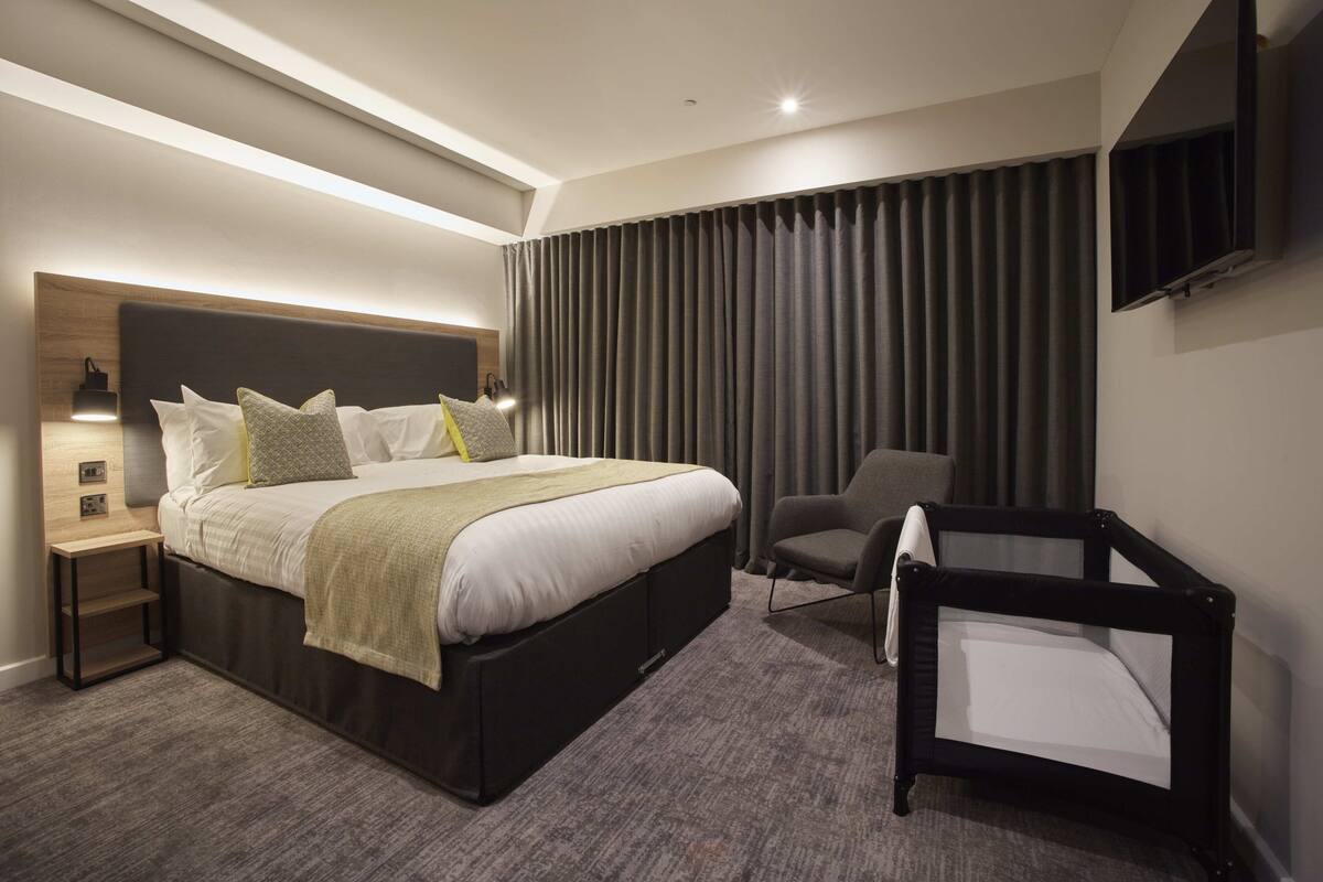 Ein modernes Hotelzimmer mit einem großen Bett, einem kleinen Schreibtisch, einem Sessel und einem an der Wand befestigten Fernseher.