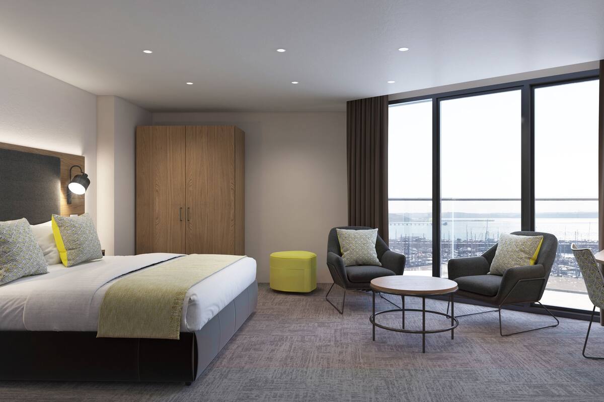 Ein modernes Hotelzimmer mit einem großen Bett, zwei Sesseln, einem kleinen Tisch und Blick auf die Stadt durch große Fenster.