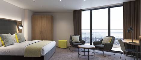 A modern hotel room with a large bed, two armchairs, a small table, and a view of the city through large windows.