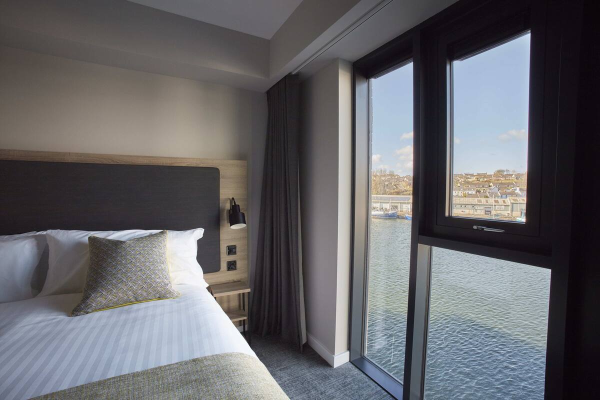 Ein Hotelzimmer mit einem großen Bett, einem Nachttisch, einer Lampe und Blick auf Wasser und Gebäude durch das Fenster.