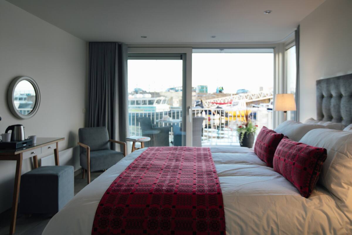 Ein Hotelzimmer mit einem großen Bett, einem Schreibtisch mit Stuhl, einem kleinen Tisch und Blick auf eine Stadtlandschaft durch das Fenster.