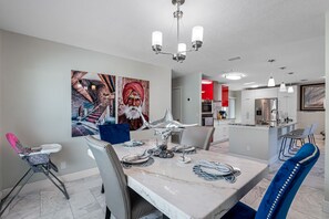 A dining area with a marble table, chairs, and a chandelier. There is a kitchen in the background with modern appliances and a red accent wall.