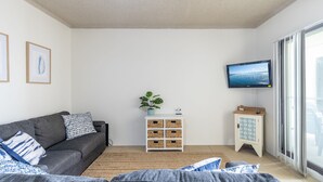 Un salon moderne avec un canapé gris, un petit meuble blanc avec des paniers, une plante en pot et un téléviseur fixé au mur.