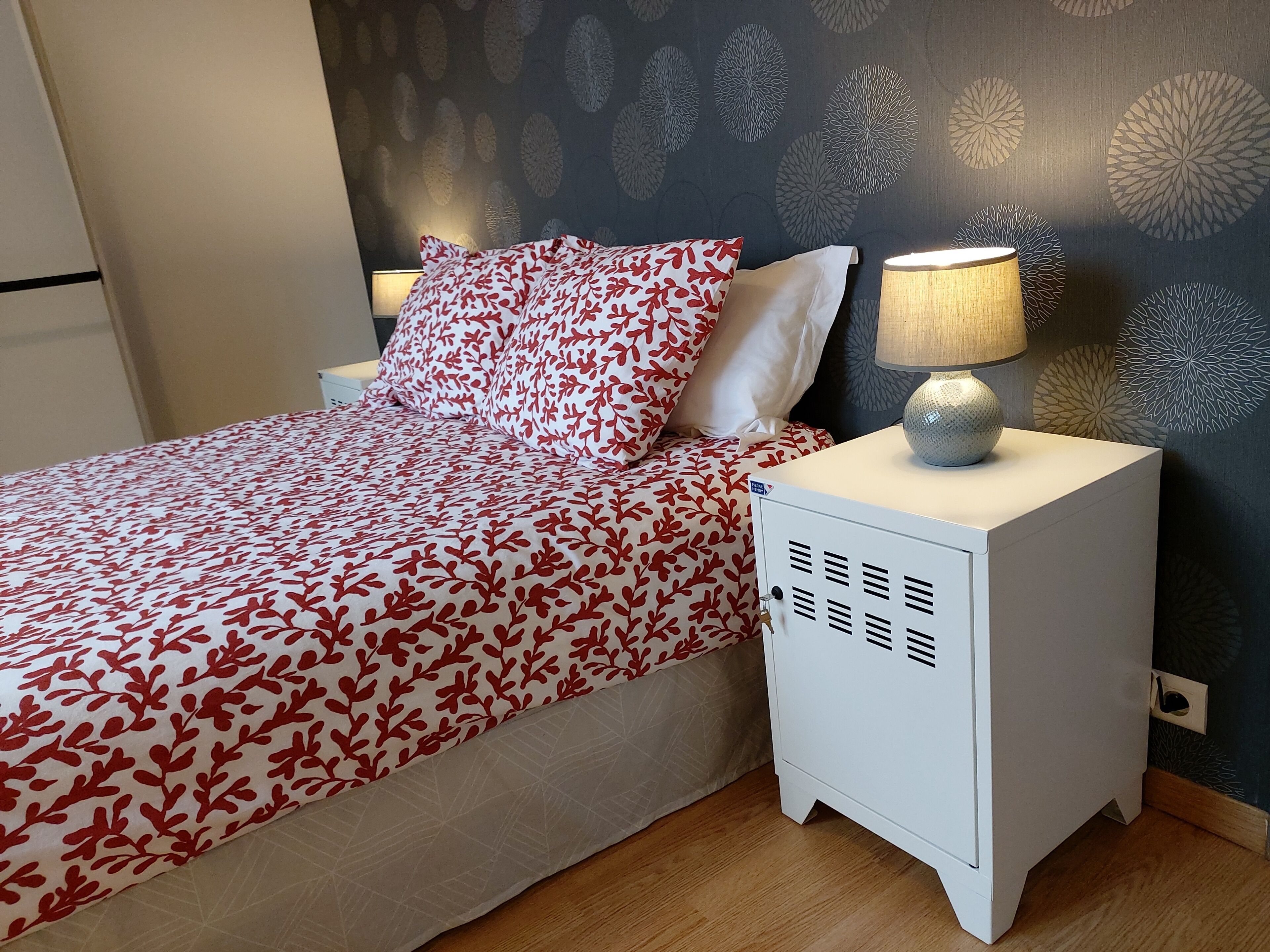Un lit bien fait, recouvert d’une couvre-lit à motifs, une lampe de chevet et une petite armoire blanche avec une serrure.