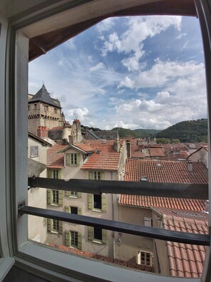 Une vue sur un paysage urbain à travers une fenêtre, avec un mélange de bâtiments traditionnels et modernes, dont certains sont coiffés de toits en tuiles rouges, et une tour imposante qui se distingue.