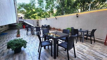 Un patio al aire libre con varias mesas y sillas, rodeado de plantas en macetas y una pared con luces fijadas.