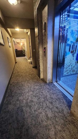 Un pasillo con pisos alfombrados, una puerta de vidrio que da al exterior y un panel digital de ascensor.