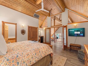 A bedroom with a large bed, wooden ceiling beams, a TV, and a staircase leading to an upper level.