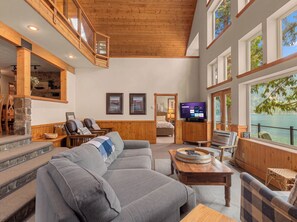 Un vaste espace de vie agrémenté d’un grand canapé gris, d’éléments décoratifs en bois, d’un téléviseur et offrant une vue sur un lac.