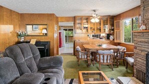 Uma sala de estar com sofá, mesa de jantar com cadeiras, área de cozinha com armários e uma geladeira, e um piano.